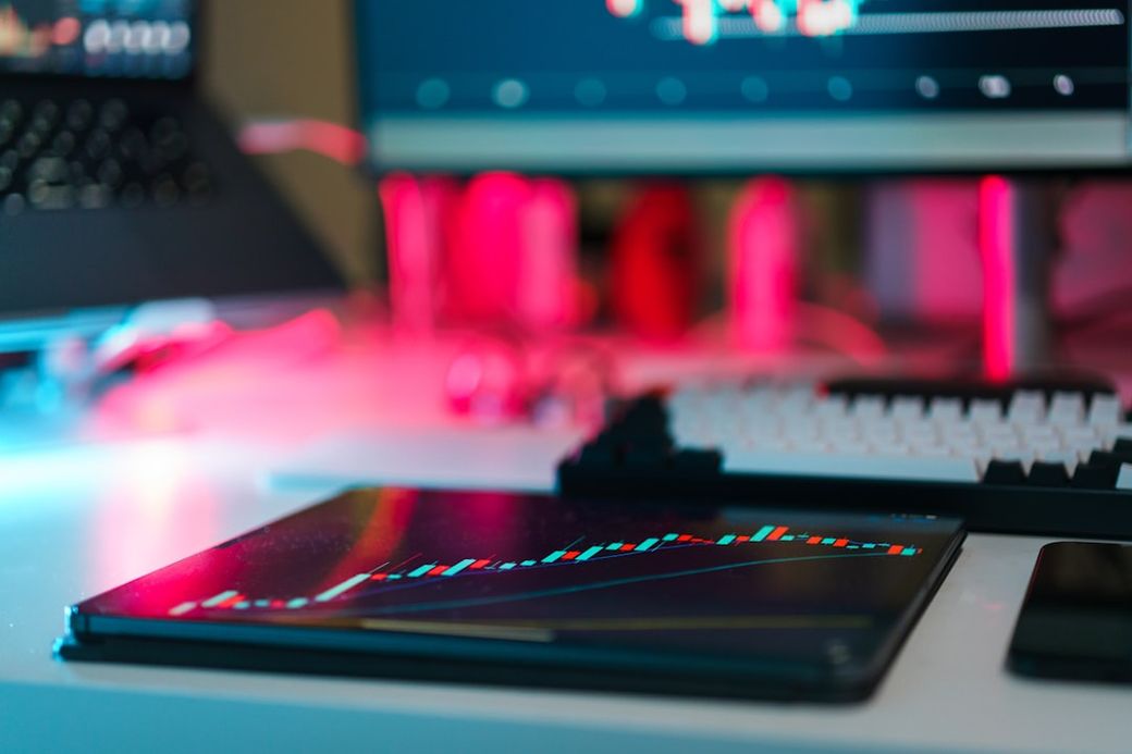 The image shows a close-up of a tablet displaying a stock market chart on a desk, with a keyboard and a laptop in the background. The scene is illuminated with vibrant lights, highlighting the modern workspace. — Credit: Photo by Jakub Żerdzicki on Unsplash