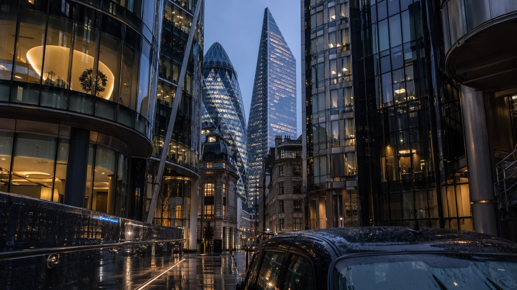 The image showcases a modern cityscape featuring sleek skyscrapers with glass facades, reflecting twilight hues. A street with a parked vehicle is visible in the foreground, highlighting the urban architecture and atmosphere.
