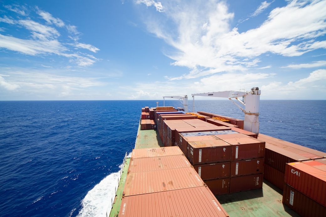 The image captures the deck of a cargo ship navigating through open ocean waters, with shipping containers stacked on one side. The sky is bright blue with scattered clouds, emphasizing the vastness of the sea. — Credit: Photo by Kurt Cotoaga on Unsplash