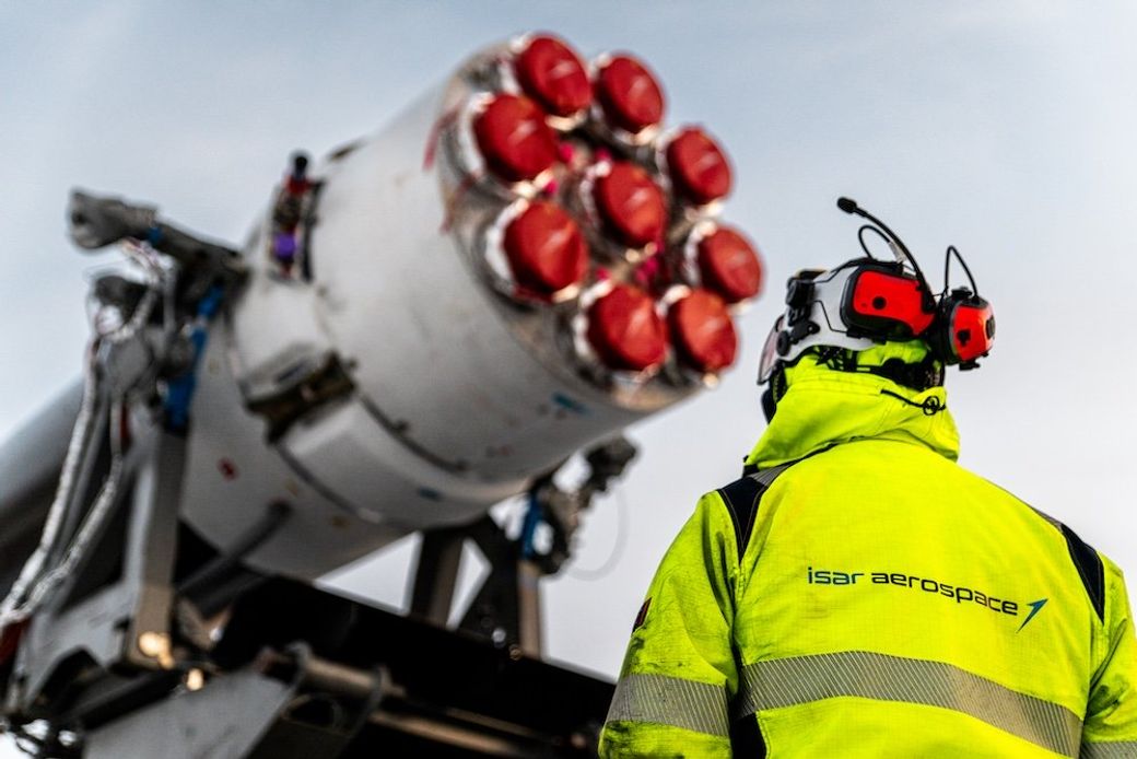 A worker in a high-visibility jacket observes a rocket's engine, which features multiple red nozzles. The setting appears to be a launch pad or aerospace facility, highlighting preparations for a possible launch.