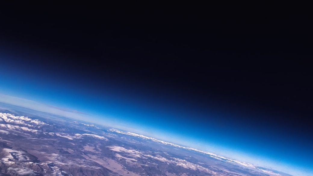 The image captures a view of Earth's curvature from space, showing the transition from the planet's surface to the dark expanse of space. Snow-capped mountains and varied terrain are visible below, juxtaposed against the gradient of the atmosphere. — Credit: Photo by ActionVance on Unsplash