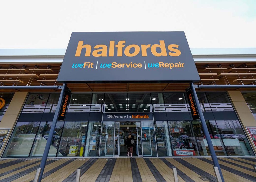 The image shows the entrance of a Halfords store, prominently displaying its branding overhead. Customers are seen entering the store, which specializes in automotive and cycling products and services.