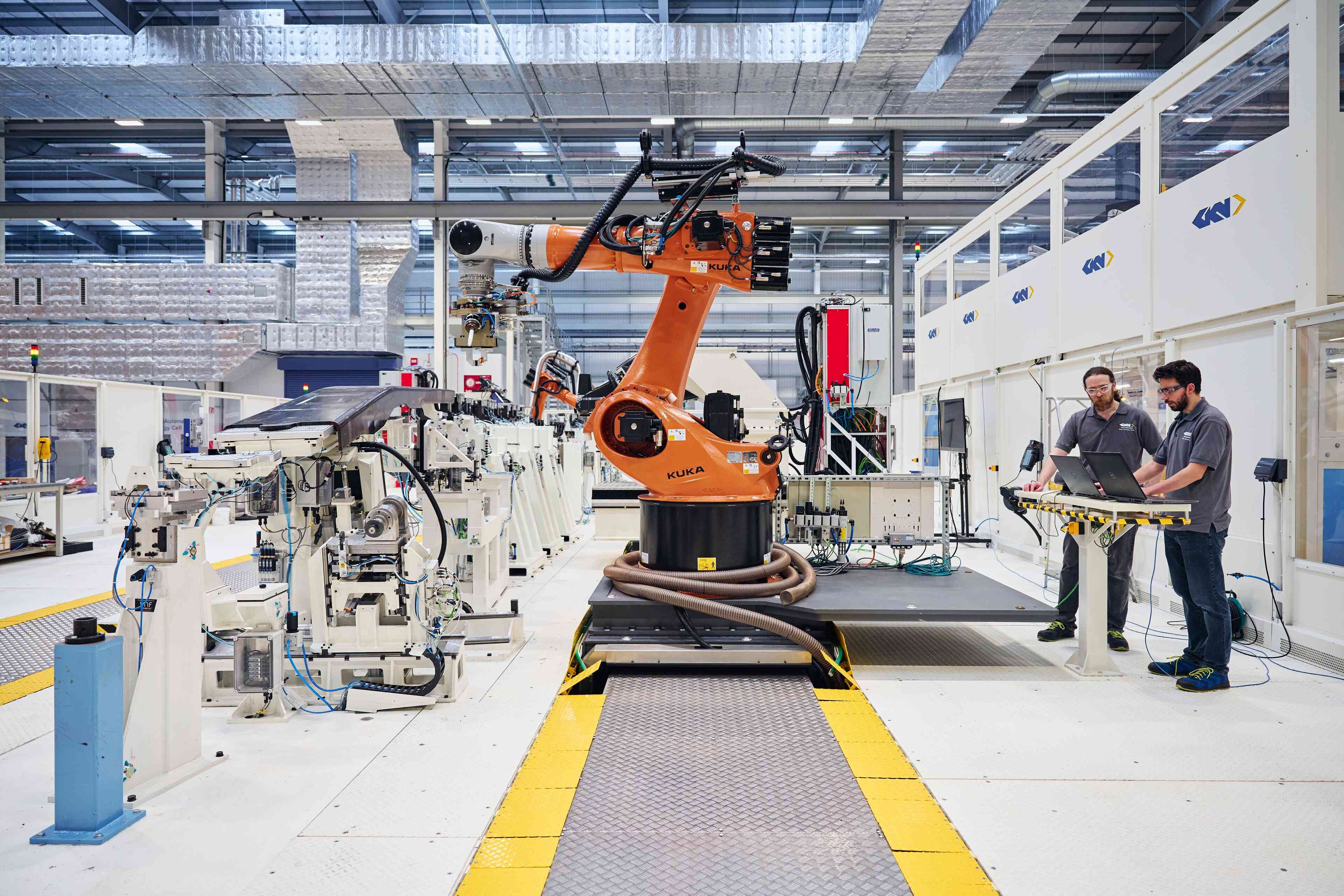 The image shows an advanced manufacturing facility with two technicians working alongside an industrial robotic arm. The robot is positioned on a production platform, highlighting automation in the manufacturing process.