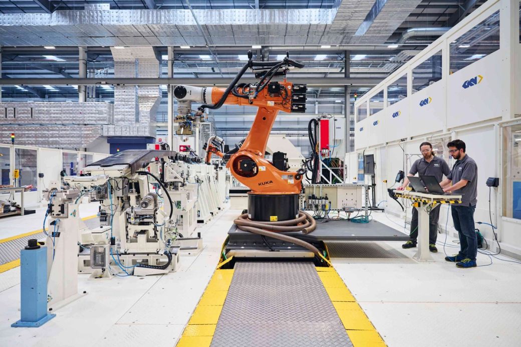 The image shows an advanced manufacturing facility with two technicians working alongside an industrial robotic arm. The robot is positioned on a production platform, highlighting automation in the manufacturing process.