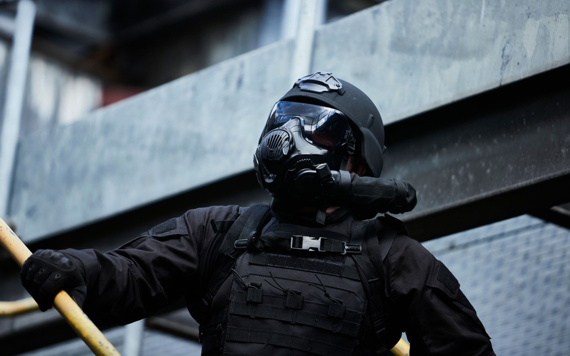 A tactical officer in full gear, including a black helmet and gas mask, is captured in an industrial setting. The figure stands against a backdrop of metal structures, suggesting a high-stakes operation.