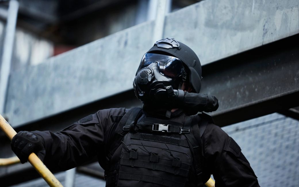 A tactical officer in full gear, including a black helmet and gas mask, is captured in an industrial setting. The figure stands against a backdrop of metal structures, suggesting a high-stakes operation.
