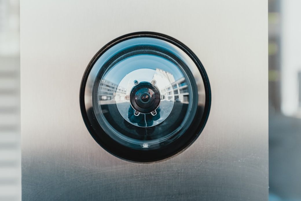 The image features a close-up view of a security camera lens mounted on a metallic surface. The reflection captured in the lens shows a blurred view of buildings and surroundings, indicating an urban environment. — Credit: Photo by Bernard Hermant on Unsplash