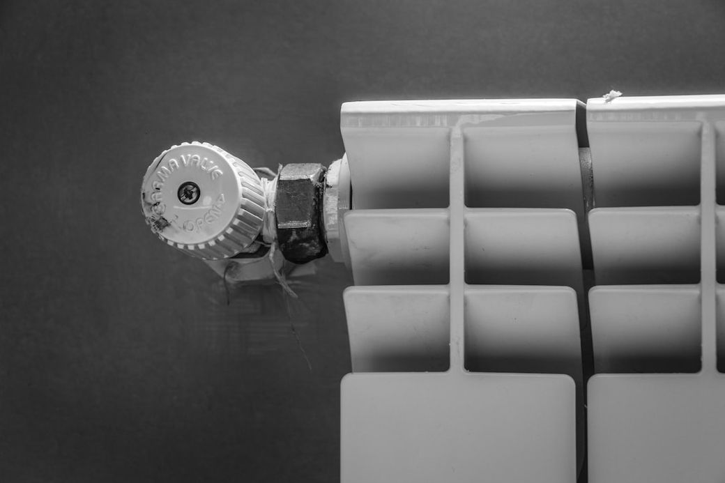 The image features a close-up view of a radiator valve and section of a radiator. The photograph is presented in black and white, highlighting the textures and details of the materials. — Credit: Photo by hessam nabavi on Unsplash
