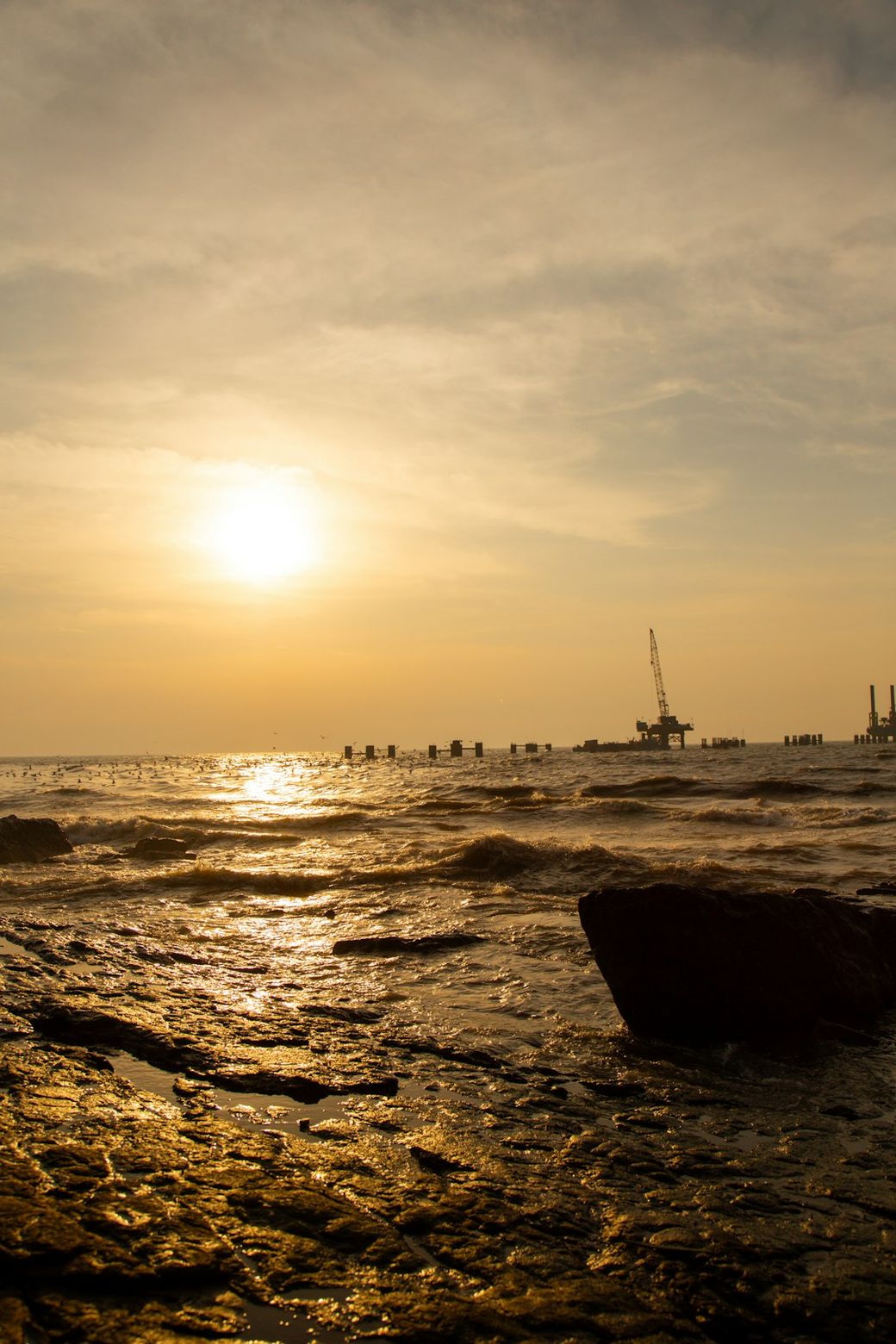 The image captures a serene coastal scene during sunset, featuring a rocky shoreline with gentle waves lapping against the rocks. In the background, an industrial structure and crane are silhouetted against the vibrant sky. — Credit: Photo by Ishaan Sen on Unsplash