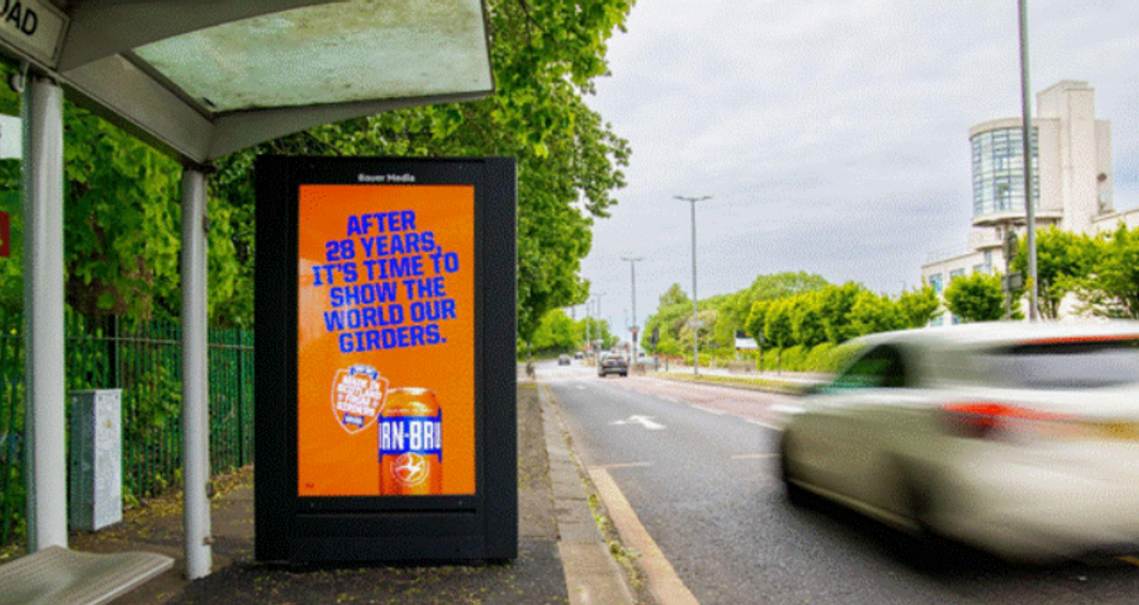 The image shows a roadside bus stop with a large advertisement featuring a beverage. The billboard reads an announcement celebrating 28 years with a call to action to show off their 'girders', while vehicles pass by on the street.