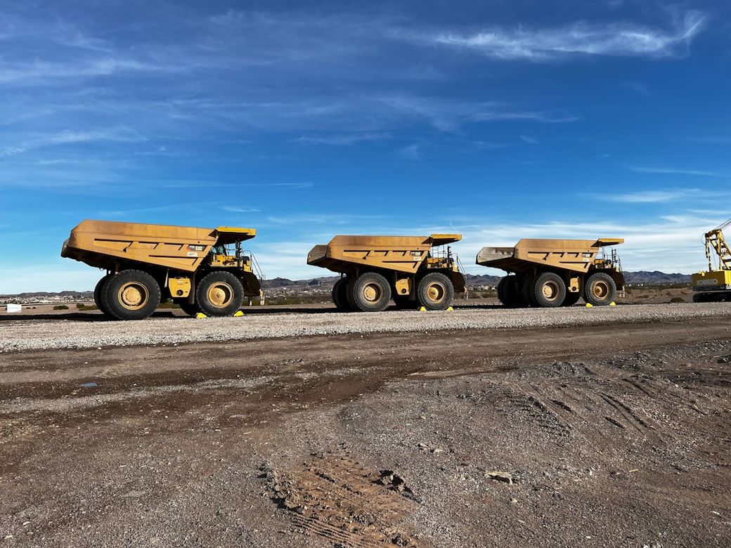 The image shows three large yellow dump trucks lined up on a gravel surface, set against a clear blue sky and distant mountains. The trucks are designed for heavy-duty transport, often seen in mining or heavy construction operations. — Credit: Photo by Boom & Bucket on Unsplash