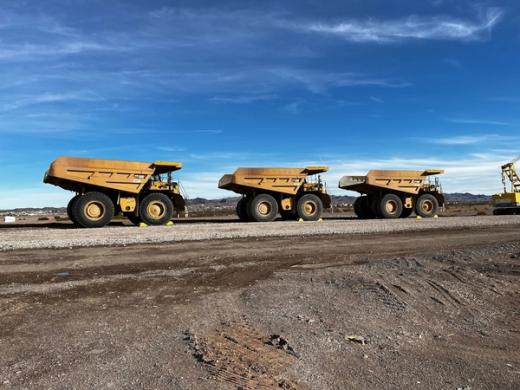 The image shows three large yellow dump trucks lined up on a gravel surface, set against a clear blue sky and distant mountains. The trucks are designed for heavy-duty transport, often seen in mining or heavy construction operations.