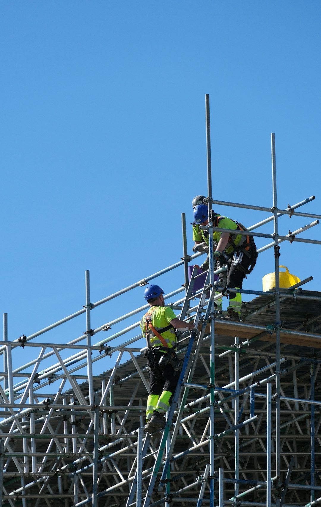 Two construction workers are seen working on a scaffolding structure against a clear blue sky. They are engaged in tasks above ground, showcasing safety gear and tools.