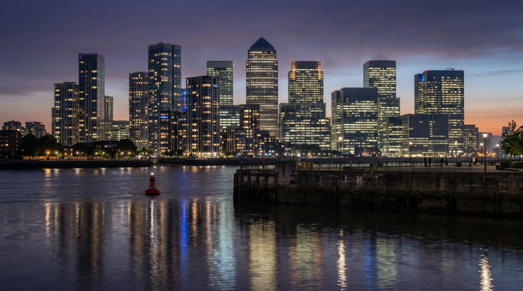 The image captures the Canary Wharf skyline at dusk, reflecting a blend of deep violet and burnt orange hues in the sky. The towers are illuminated, revealing a striking contrast of warm and cool light against the darkened water of the Thames, while a red navigation buoy adds a pop of color and scale to the foreground.