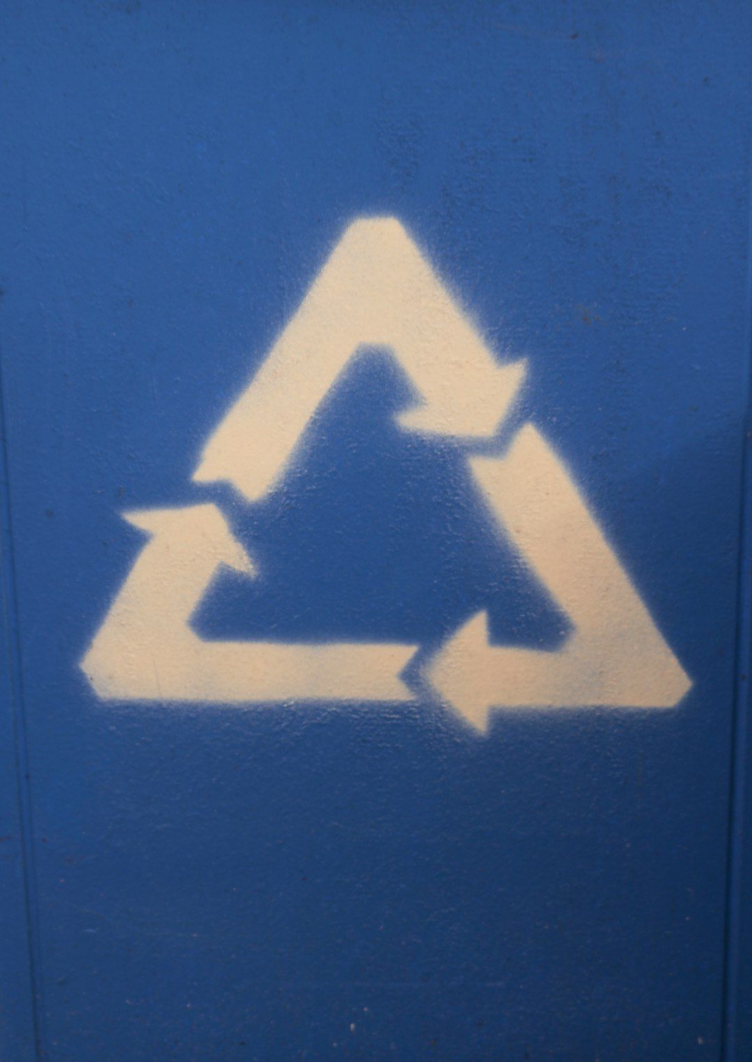 The image depicts a stenciled recycling symbol painted on a blue surface. The symbol consists of three arrows forming a triangle, representing the recycling process.