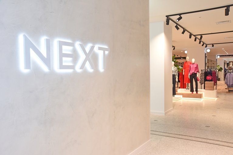 The image shows the interior of a retail store featuring clothing displays. The illuminated sign of the brand 'NEXT' is prominently visible on the left wall.