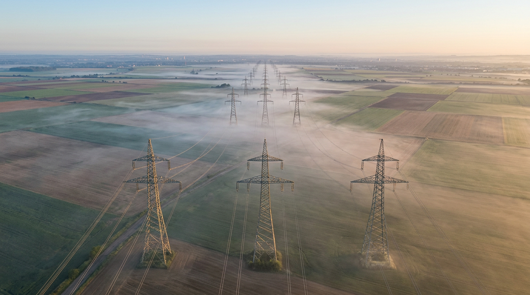 The image captures an abstract aerial view of electricity pylons evenly spaced across flat agricultural land, extending toward the horizon. The pylons rise above early morning mist, their geometric forms contrasted by the organic patchwork of fields below, highlighted by golden-hour light and creating a rhythmic pattern across the landscape.