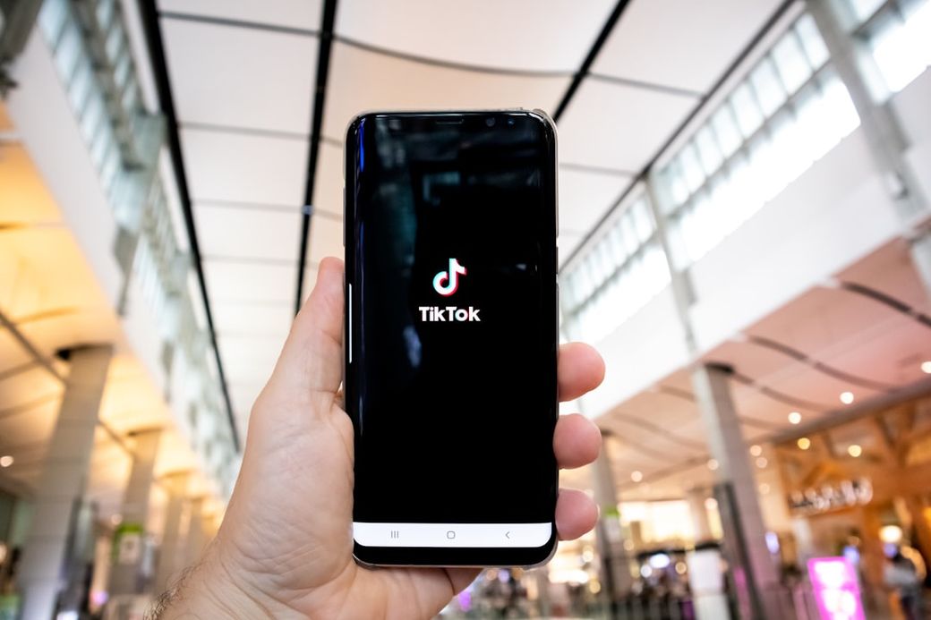 A person is holding a smartphone displaying the TikTok logo indoors, likely in a public space such as a shopping center or airport. The environment features modern architecture with large windows and a spacious layout. — Credit: Photo by Olivier Bergeron on Unsplash