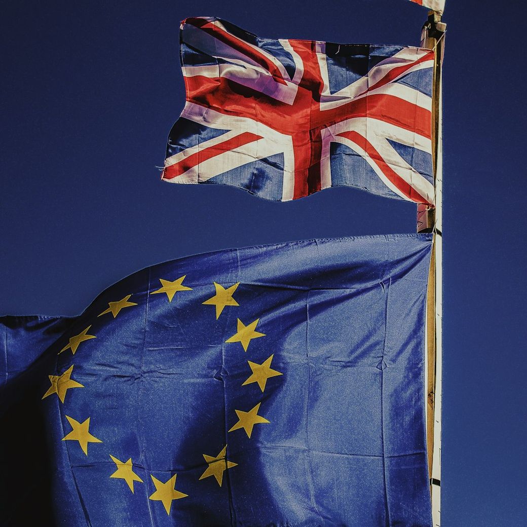 The image features the flags of the United Kingdom and the European Union flying side by side against a blue background. The Union Jack and the EU flag are prominently displayed, symbolizing the political relationship between the UK and the EU. — Credit: Photo by Rocco Dipoppa on Unsplash
