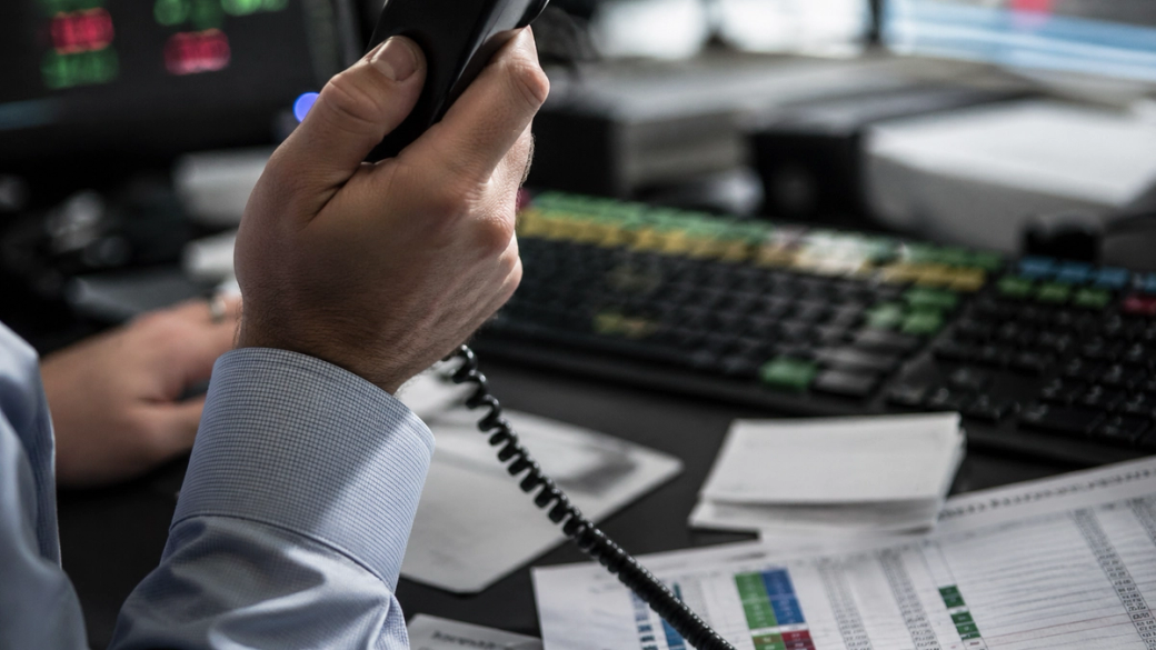 A close-up shot of a person holding a phone, engaged in what appears to be a financial discussion. The workspace features a computer with stock market data on screen and various financial documents scattered on the desk.