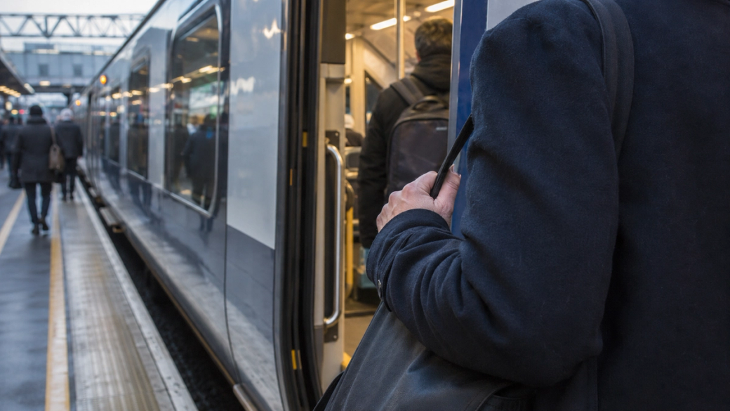 A commuter is seen waiting at a train station, standing near the doorway of a train that has just arrived. The train platform is wet, indicating recent rainfall, and passengers are boarding while others are waiting nearby.