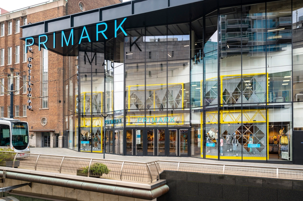 The image depicts the storefront of a Primark retail store, characterized by its large glass facade and modern architecture. The entrance features bright yellow accents and displays a range of clothing visible through the windows.