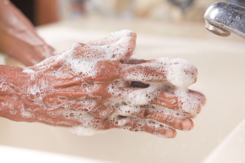 The image depicts a close-up of hands washing with soap under running water. The hands are heavily soaped and show signs of aging, indicating an emphasis on hygiene and cleanliness. — Credit: Photo by Towfiqu barbhuiya on Unsplash