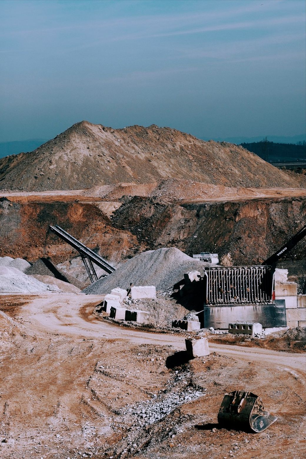 The image shows a mining site characterized by large piles of earth and gravel, with machinery used for material processing prominently in the foreground. The landscape features a mix of crushed stone and gravel, indicating ongoing excavation and resource extraction activities. — Credit: Photo by David Hellmann on Unsplash