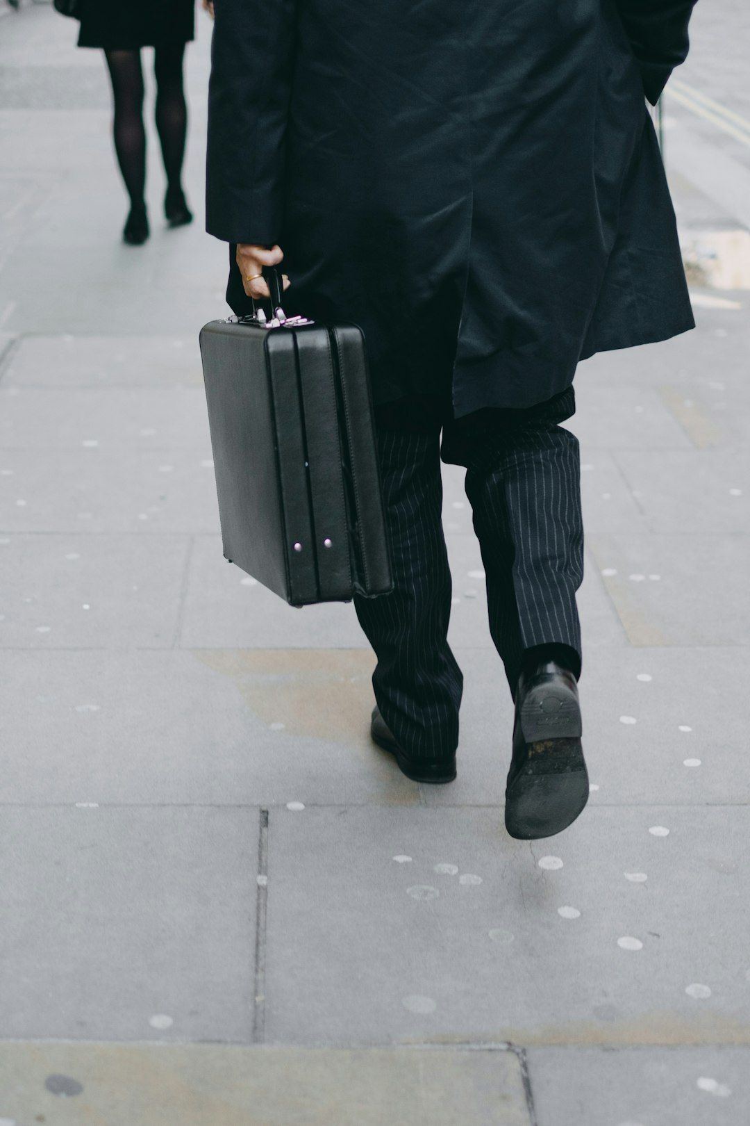 A man in formal attire carrying a briefcase walks down a city sidewalk. His pinstripe pants and long coat are visible, suggesting a professional setting.