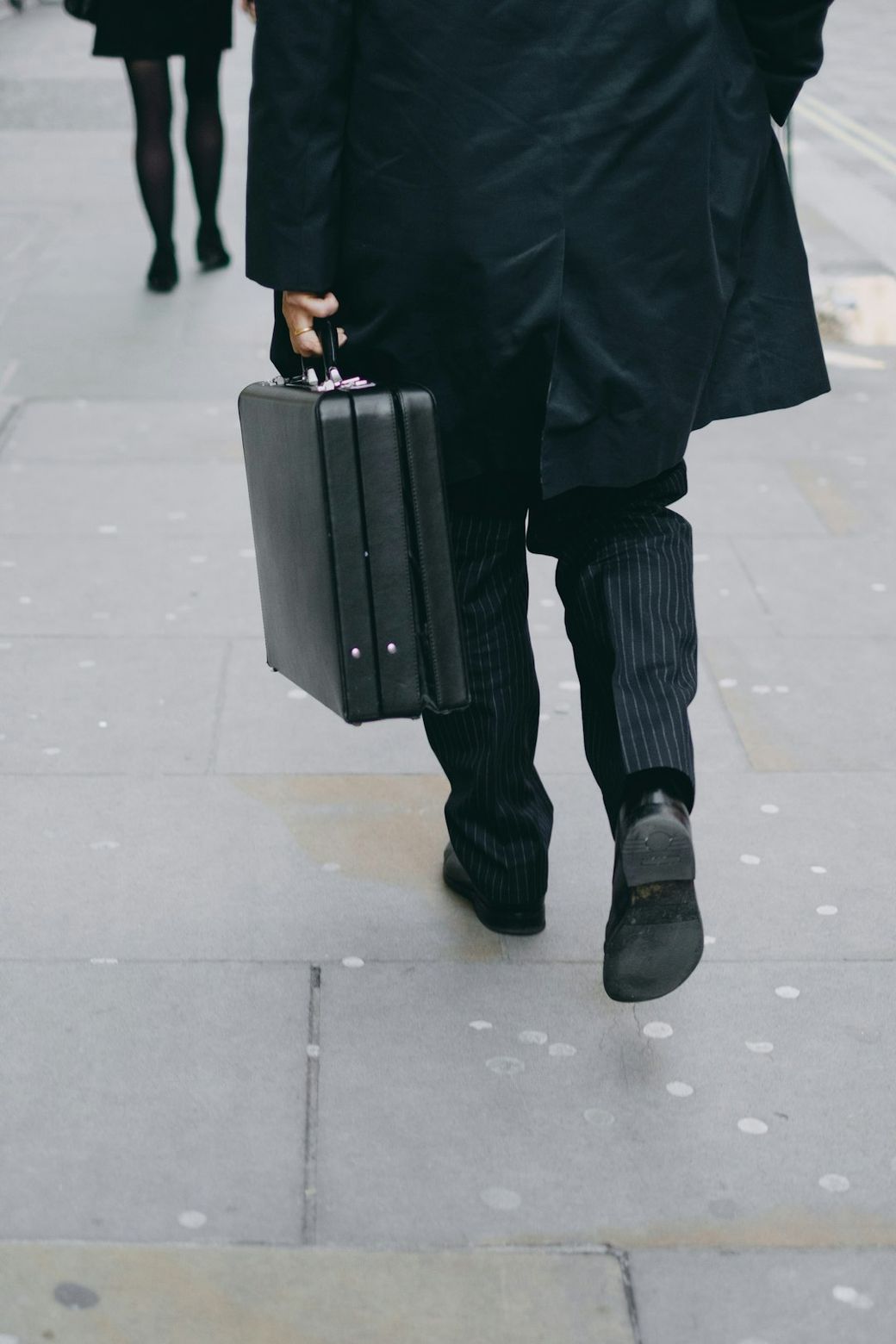 A man in formal attire carrying a briefcase walks down a city sidewalk. His pinstripe pants and long coat are visible, suggesting a professional setting. — Credit: Photo by Craig  Whitehead on Unsplash