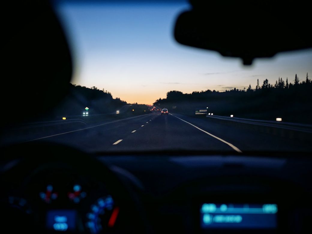 The image captures a nighttime driving scene from the perspective of a driver inside a car. The road extends ahead, with faint lights of oncoming vehicles and a gradient sky transitioning from dusk to night. — Credit: Photo by Patrick Tomasso on Unsplash