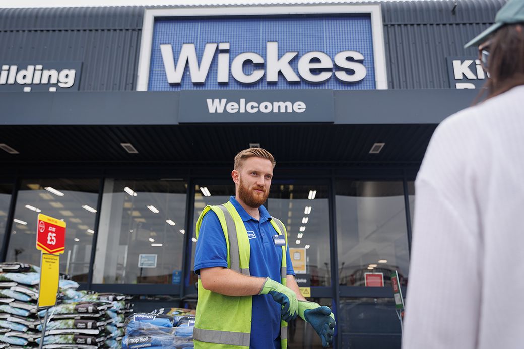 A worker stands outside a Wickes home improvement store, wearing a safety vest and gloves. The store's sign is prominently displayed, welcoming customers.