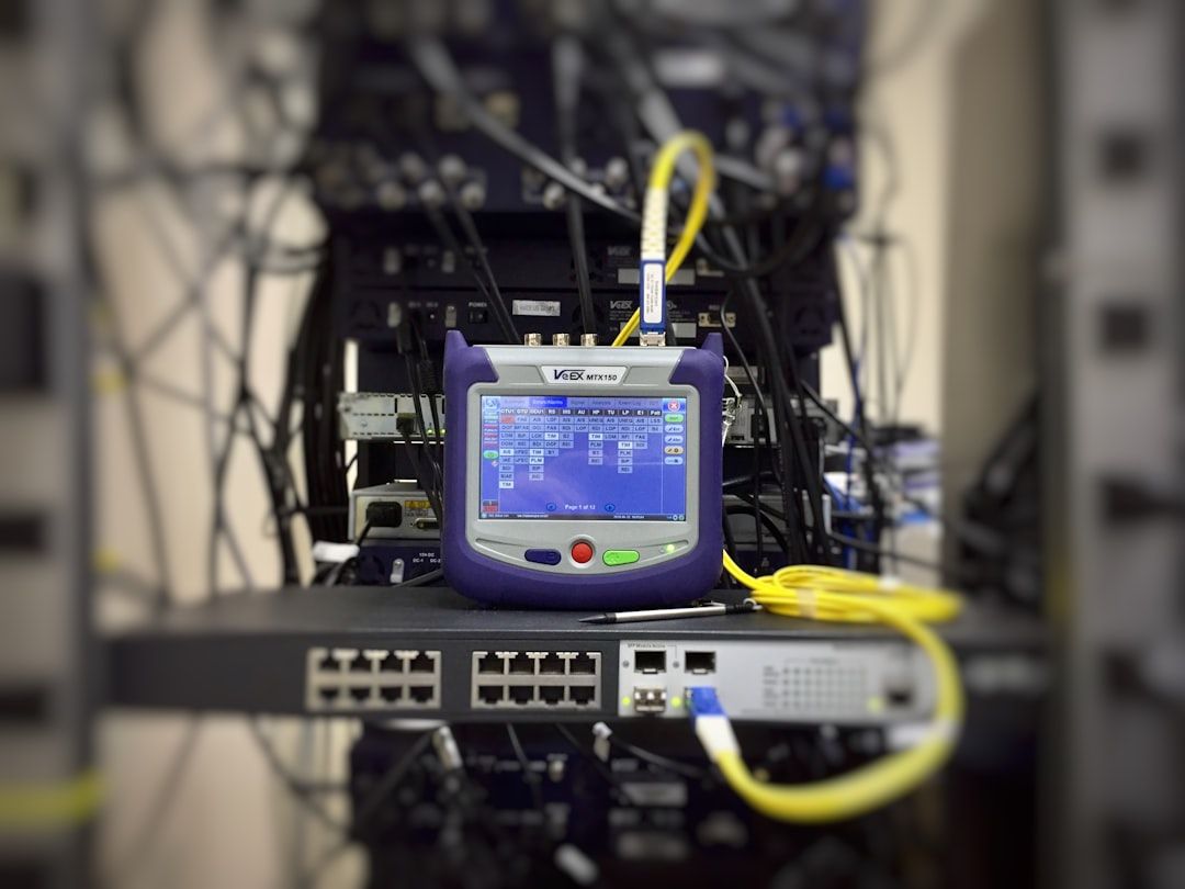 The image shows a network testing device positioned in front of a network rack filled with cables and equipment. The device's screen displays various connection metrics and diagnostics, indicating it is used for analyzing network performance.