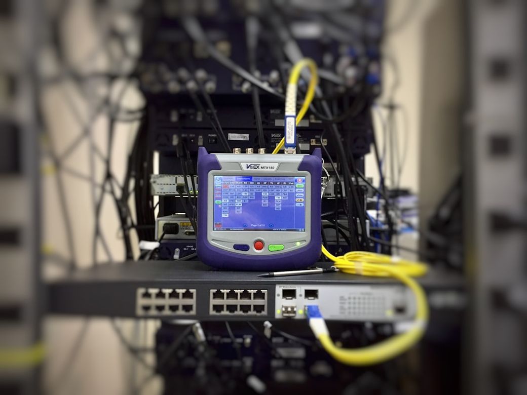 The image shows a network testing device positioned in front of a network rack filled with cables and equipment. The device's screen displays various connection metrics and diagnostics, indicating it is used for analyzing network performance. — Credit: Photo by Ildefonso Polo on Unsplash