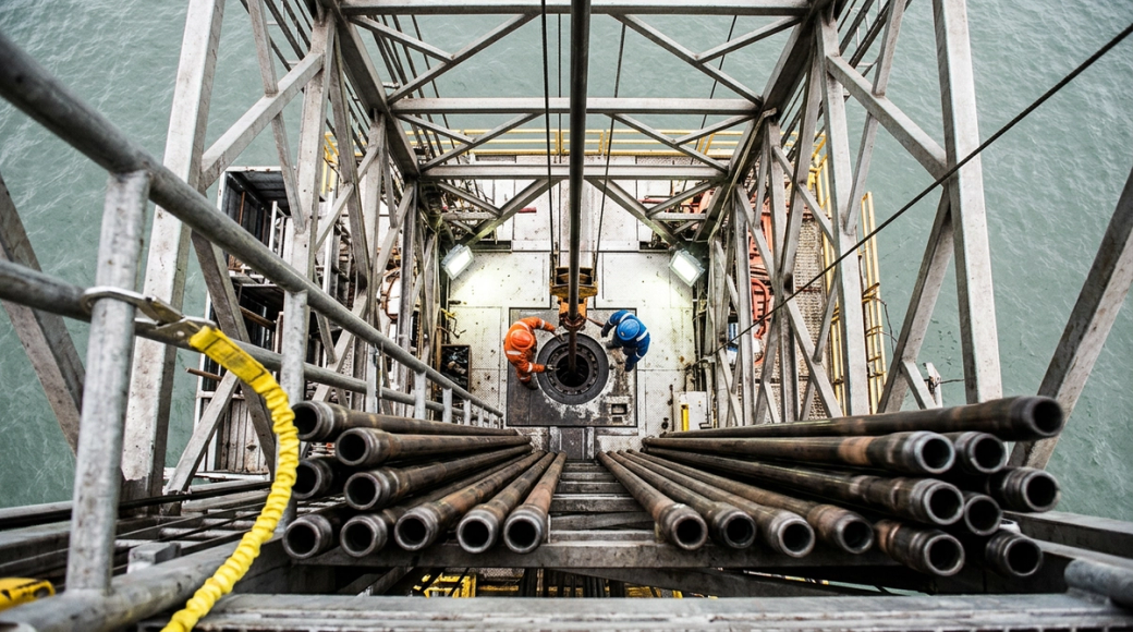 The image captures a downward view from a monkey board on an oil drilling rig, showcasing the drill floor below. Two figures in hard hats and coveralls are positioned by the rotary table, which is central to the scene, surrounded by a complex lattice of steel structures. The composition emphasizes the height and precariousness of the platform, with rows of racked pipe and a distant view of the sea below.