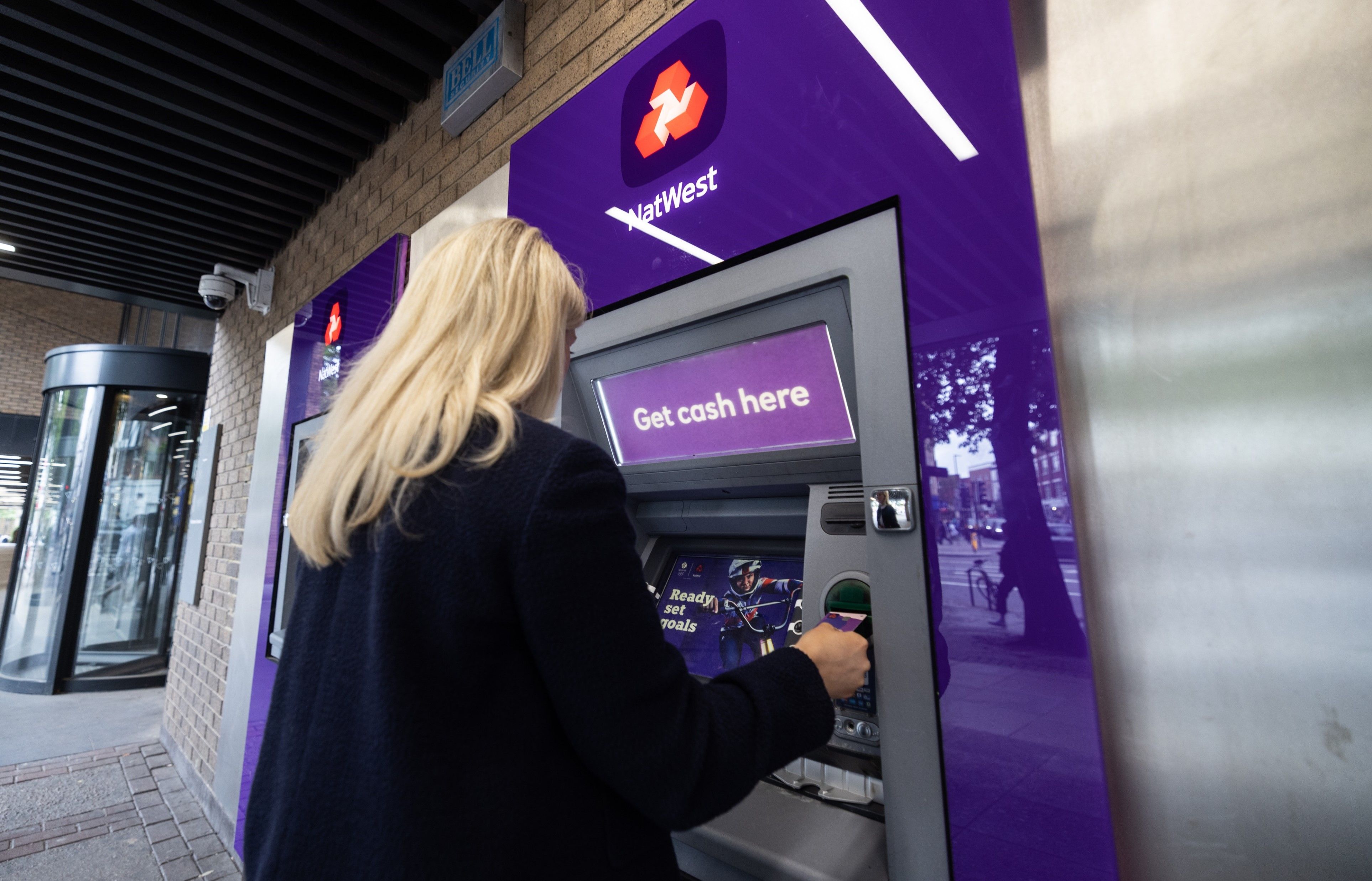 A person is using an ATM machine outside a bank. The ATM is branded with the logo of NatWest and features bright purple signage. The individual is engaged in a transaction at the machine.