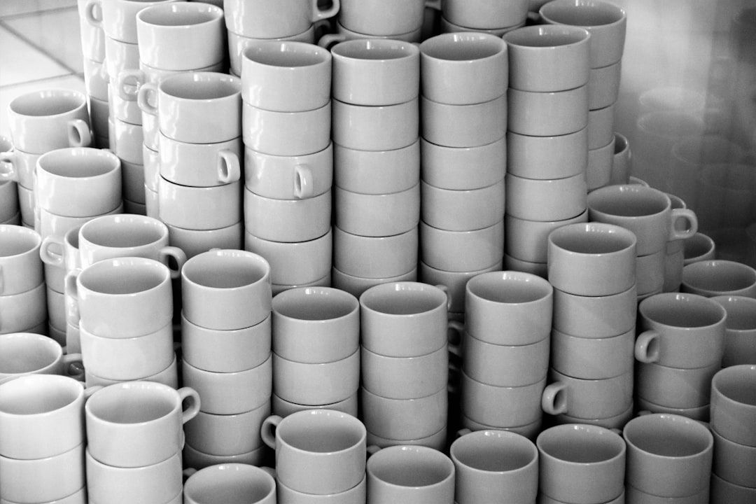 The image depicts a large stack of white ceramic cups arranged in a triangular formation. The monochrome color scheme emphasizes the shapes and textures of the cups, creating a visually striking pattern.