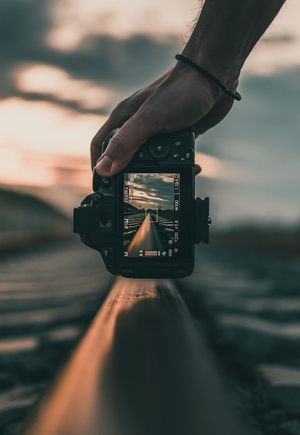 The image features a hand holding a camera aimed at railroad tracks, capturing a reflection of the landscape in the camera screen. The scene is set during sunset, with dramatic clouds and a golden light illuminating the tracks. — Credit: Photo by Fotis Fotopoulos on Unsplash