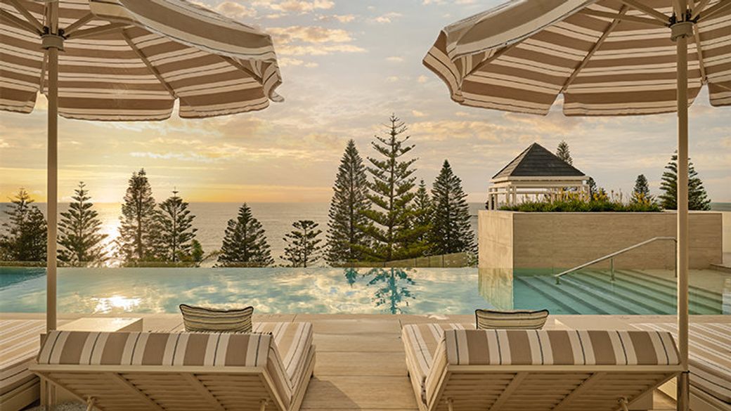 The image showcases a serene resort poolside setting at sunset, featuring loungers with striped umbrellas and a backdrop of lush evergreen trees and the ocean. The tranquil scene evokes a sense of relaxation and luxury.