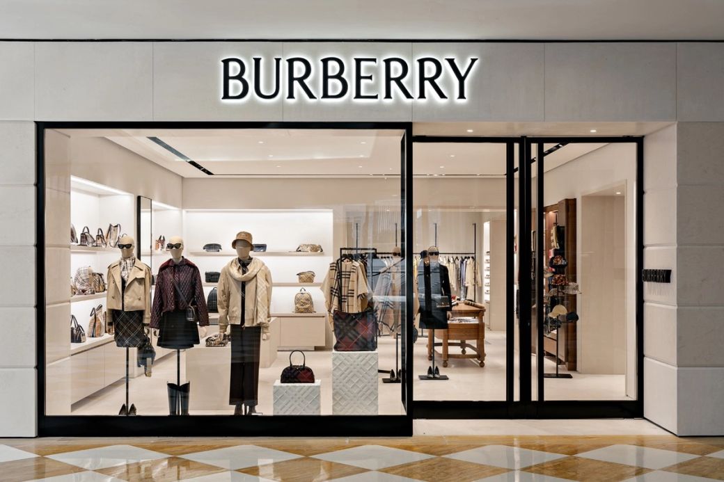 The image shows a Burberry retail store facade featuring mannequins dressed in various designer outfits. The store is well-lit with an organized display of luxury bags and accessories visible through the glass.