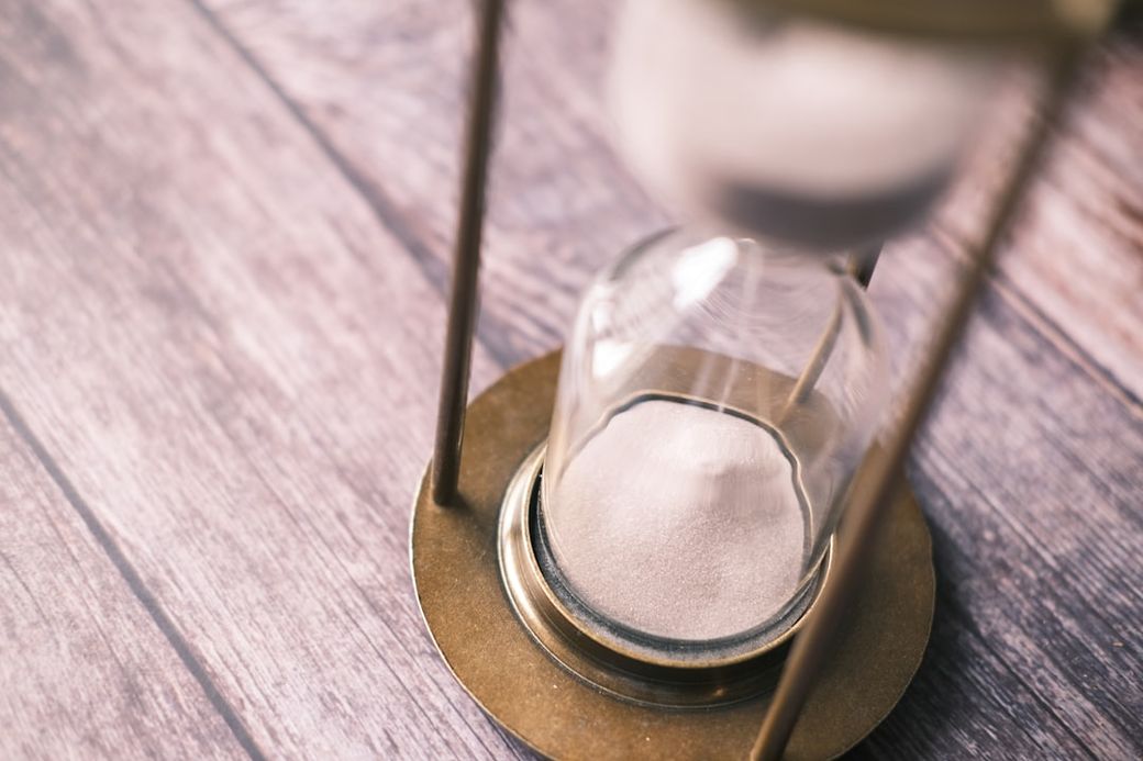 The image features a close-up of an hourglass, with sand gently flowing from the upper bulb to the lower bulb. The hourglass is set against a wooden surface, creating a warm and rustic background. — Credit: Photo by Towfiqu barbhuiya on Unsplash
