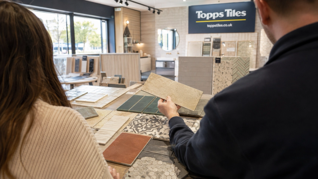 A person is examining tile samples in a showroom setting, with various tile designs displayed on tables. The interior is well-lit, showcasing a modern design aesthetic.