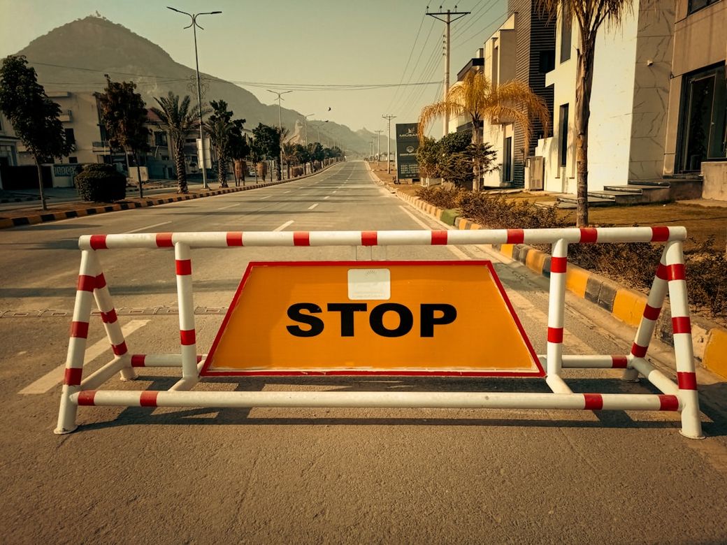 The image shows a roadblock with a prominent 'STOP' sign in the foreground. The road appears deserted and lined with palm trees, set against a backdrop of a mountainside. — Credit: Photo by Fahim Junaid on Unsplash