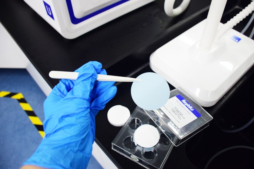 A laboratory setting where a person wearing a blue glove holds a circular blue sample on a spatula. Nearby, there are circular containers and a labeled box on a work surface, suggesting an experiment or testing process.