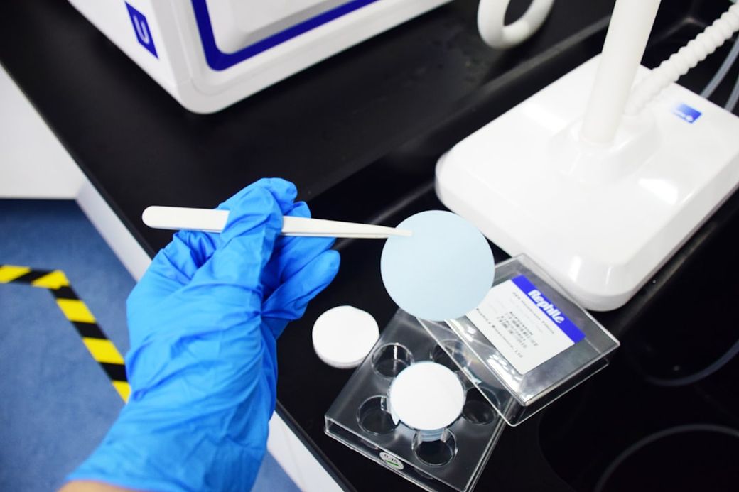A laboratory setting where a person wearing a blue glove holds a circular blue sample on a spatula. Nearby, there are circular containers and a labeled box on a work surface, suggesting an experiment or testing process. — Credit: Photo by RephiLe water on Unsplash