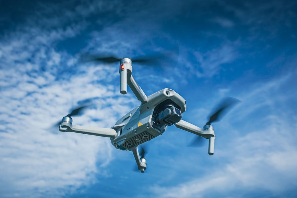 A drone is flying against a backdrop of a blue sky with white clouds. The image captures the drone mid-air, showcasing its four propellers in motion. — Credit: Photo by Harrison Kugler on Unsplash