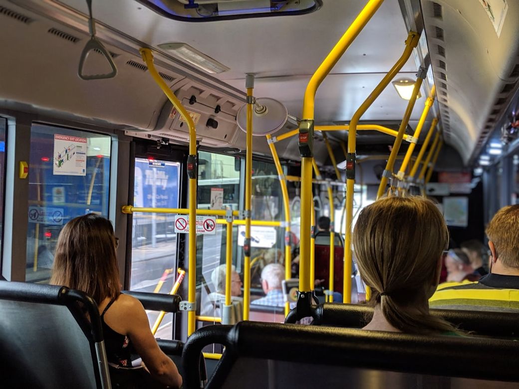 The image shows the interior of a city bus with passengers seated and standing. Yellow poles support the bus's structure, and the scene outside the bus is partially visible through the windows. — Credit: Photo by Mitchell Johnson on Unsplash