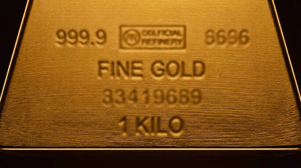 The image presents a close-up view of a gold bar, emphasizing its smooth surface and the subtle linear grain impressions from the mold. The assay marks and serial characters are softly blended into the background, enhancing the bar’s monolithic appearance while the warm lighting enriches the gold’s hue.