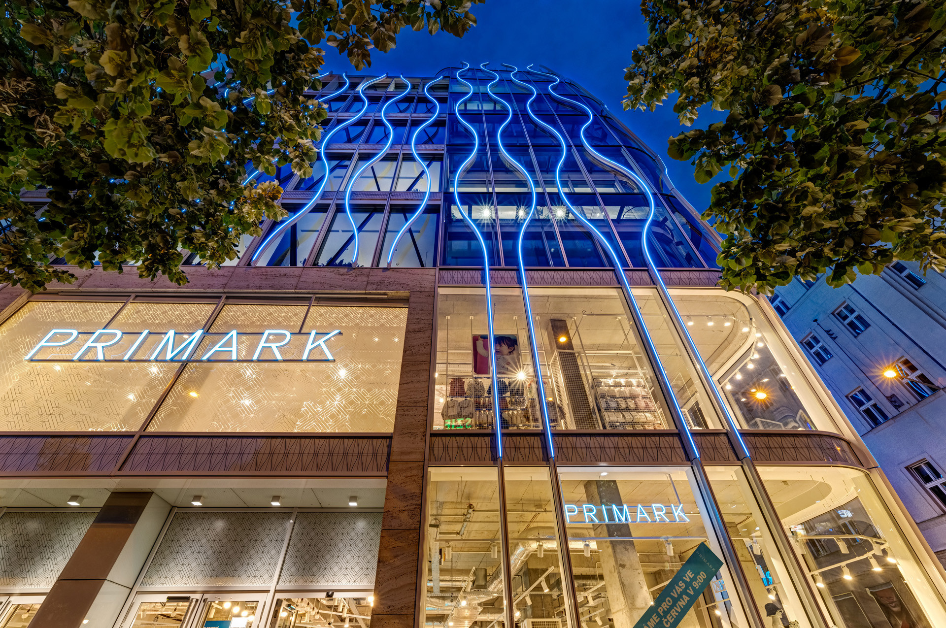 The image depicts the exterior of a Primark store, featuring illuminated signage and a modern architectural design. The building is showcased at dusk with blue lighting highlighting its unique structure.
