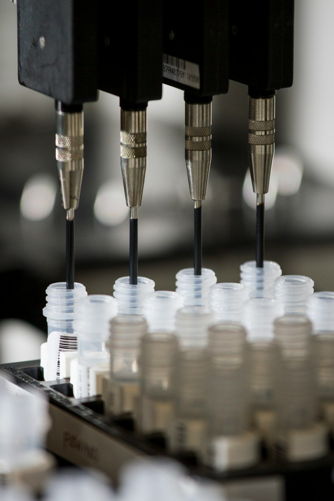 The image shows a close-up of a laboratory apparatus that is equipped with multiple precision dispensing tips. These tips are positioned above a tray filled with sample vials, suggesting a process related to automated liquid handling or chemical analysis.
